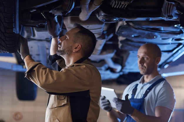 Photo portrait of two mechanics standing under car on lift while inspecting vehicle in auto repair workshop, copy space