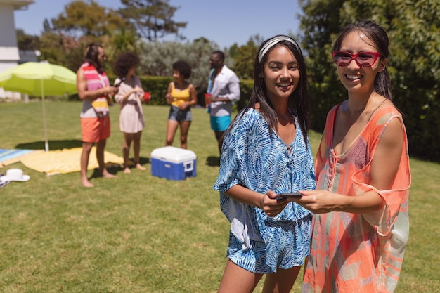 Portrait of two mixed race female friends looking at camera and smiling at a pool party. Hanging out and relaxing outdoors in summer.