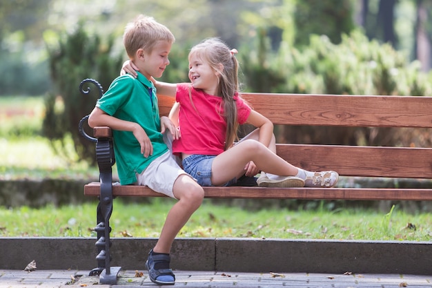 Portrait of two pretty cute children boy and girl having fun time on a bench in summer park outdoors.