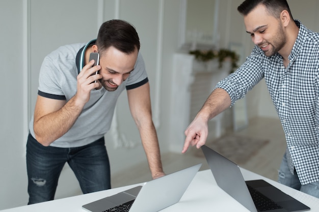 Portrait of two self-confident businessman working at laptop. One man talking on smartphone when another guy pointing with finger on screen.