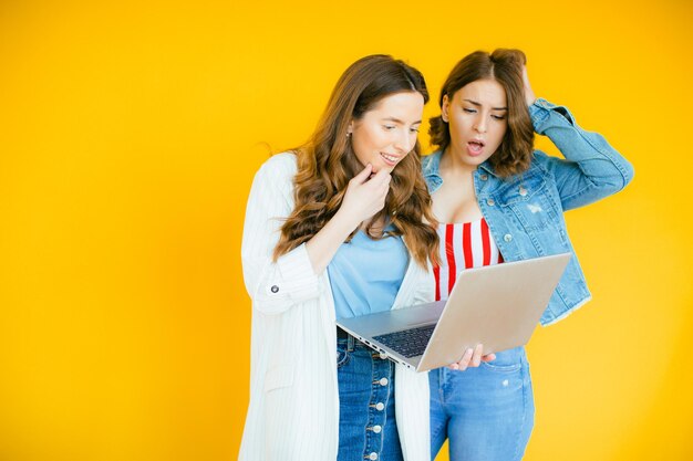 Portrait of two shocked young blackhead girls pointing finger at laptop computer on yellow