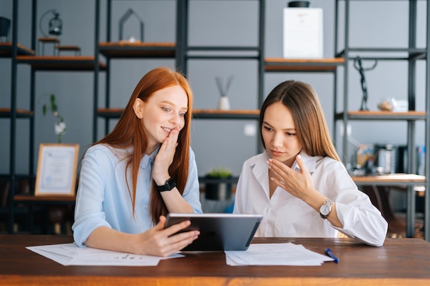 Portrait of two shocked young business women working using digital tablet at meeting desk with job documents at office. Business female colleagues using touchscreen computer for project discussion