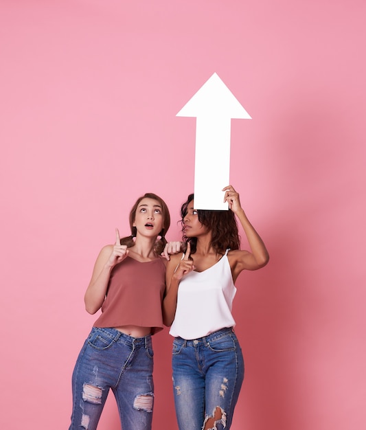 Portrait of two shocked young woman standing with her arrow pointing at copy space isolated over pink .