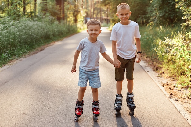 Portrait of two smiling boys brothers wearing casual styles t s-shirts and shorts rollerblading on beautiful nature on road, kids in roller skates looking at camera, spending free time in active way.