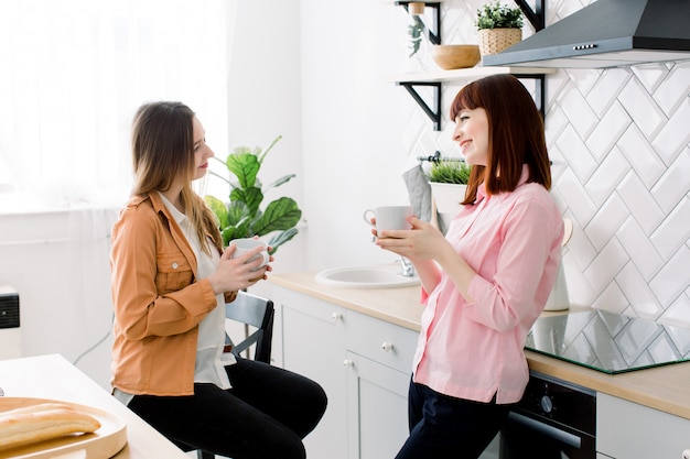 Portrait of two smiling women holding cups of coffee at home in the kitchen