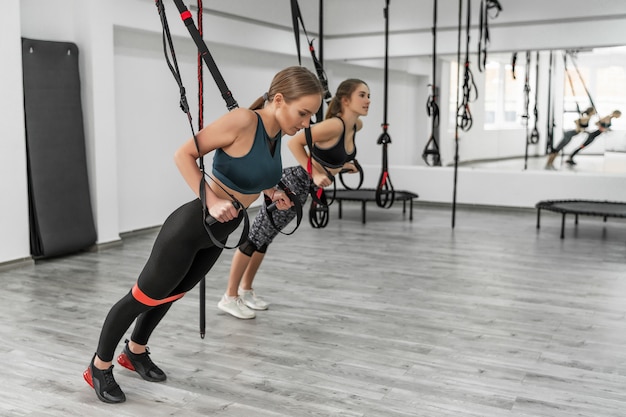 Portrait of two young beautiful fit women in sportswear training arms with trx fitness straps in the gym