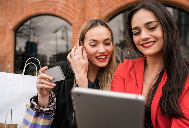 Portrait of two young friends shopping online with credit card and digital tablet while sitting outdoors. Friendship and lifestyle concept.