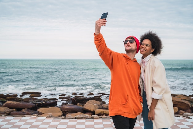 Portrait of two young friends spending good time together and taking a selfie with smartphone outdoors on the sea.