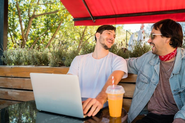 Portrait of two young friends using a laptop while sitting outdoors at coffee shop. Friendship and technology concept.