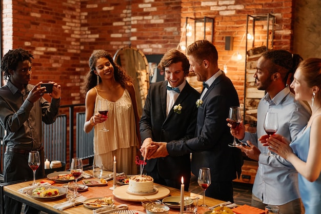 Portrait of two young men cutting cake together during wedding reception same sex marriage