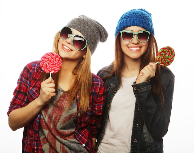 Portrait of two young pretty hipster girls wearing  hats and sunglasses holding candys. Studio portrait of two cheerful best friends having fun and making funny faces.