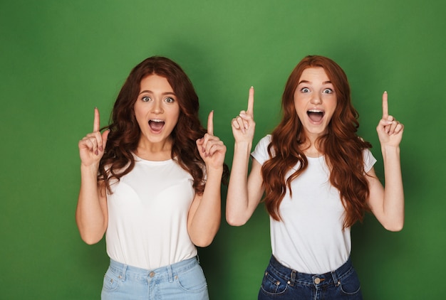 Portrait of two young redhead women 20s in white t-shirts smiling and pointing fingers upward in excitement, isolated over green background