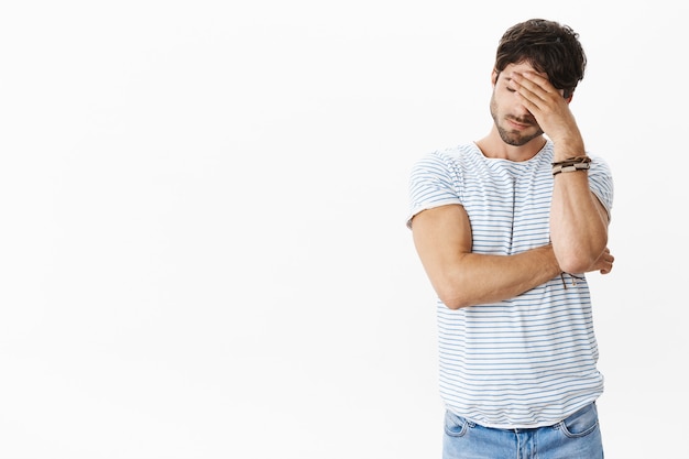 Photo portrait of uneasy exhausted and drained handsome young man in striped t-shirt closing eyes making facepalm gesture with hand on palm, covering face as feeling low