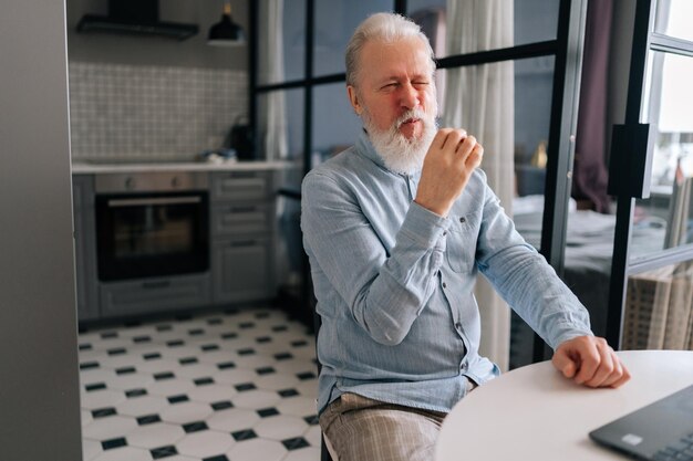 Portrait of unhappy aged grayhaired man eating unappetizing burger at kitchen home Hungry older bearded disappointed with taste of bad quality cheeseburger sitting at table and working on laptop