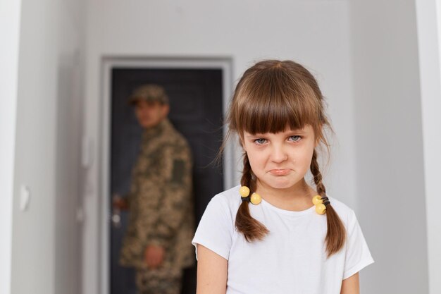 Portrait of unhappy little girl wearing white t shirt standing pout lips looking at camera being upset her father military going to war or army blurred man standing at the door