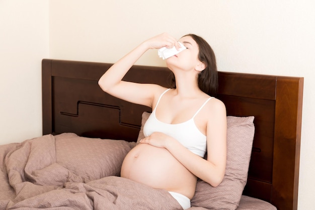 Portrait of unhappy pregnant woman leaning on pillows on bed and blowing her nose into tissue