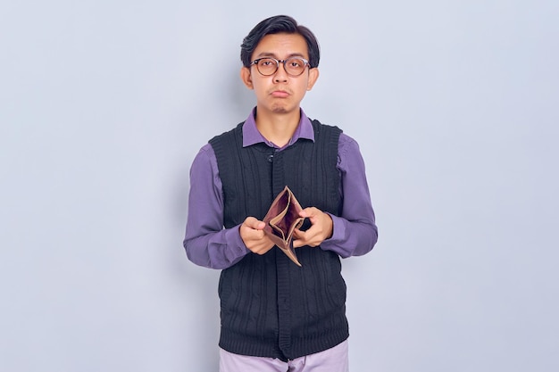 Portrait of unhappy young Asian man wearing shirt and vest showing empty wallet isolated on white background