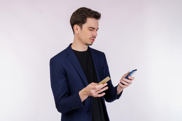 Портрет несчастного молодого бизнесмена, стоящего с мобильным телефоном и держащего кредитную банковскую карту изолированной на белом фоне студии