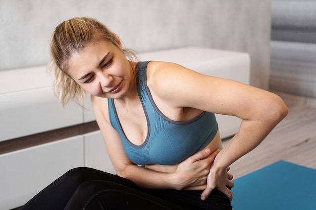 Ritratto di infelice giovane donna seduta sul materassino yoga, toccando la schiena dopo l'allenamento, soffre di mal di schiena, sensazione di dolore, vista laterale