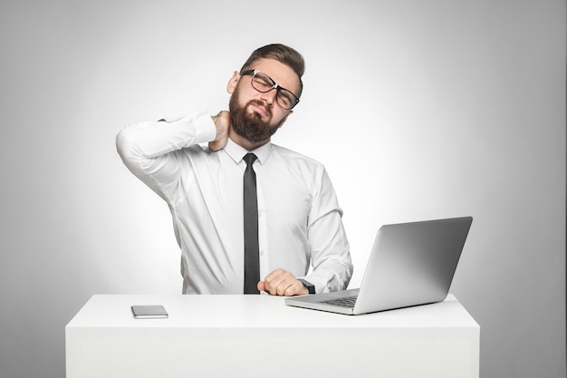 Ritratto di malsano sconvolto giovane capo stanco in camicia bianca e cravatta nera sono seduti in ufficio e hanno un forte dolore al collo, tenendo la mano sul collo. studio girato, isolato, sfondo grigio, indoor