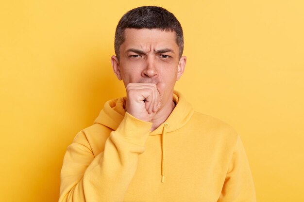 Portrait of unhealthy man wearing casual style hoodie posing isolated over yellow background coughing catches cold having high temperature having influenza symptom