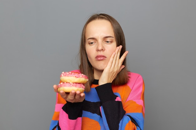 Portrait of unhealthy sad Caucasian woman wearing sweater feels pain in her teeth after eating tasty donuts touching her cheek has terribe ache posing isolated over gray background