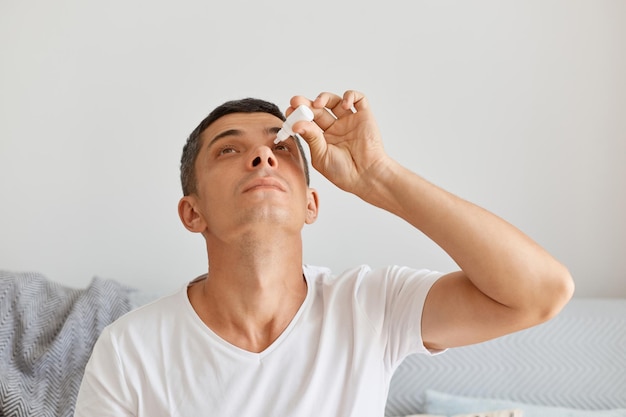 Portrait of unhealthy young adult male with dark hair wearing white t shirt, sitting on comfortable sofa in light living room, dripping eye drops to eliminate the ophthalmic problem.