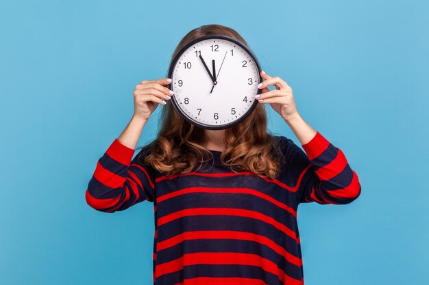 Portrait of unknown anonymous woman wearing striped casual style sweater hiding her face time management schedule and meeting appointment Indoor studio shot isolated on blue background