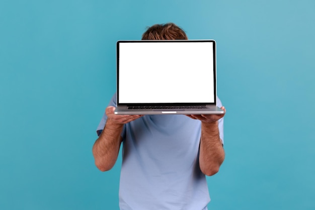 portrait of unknown man hiding face behind laptop with white empty display and presenting new applications presenting area for promotion indoor studio shot isolated on blue background