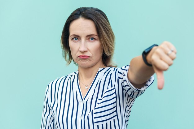 Portrait of unsatisfied sensual lady with thumbs down and white shirt against light blue background