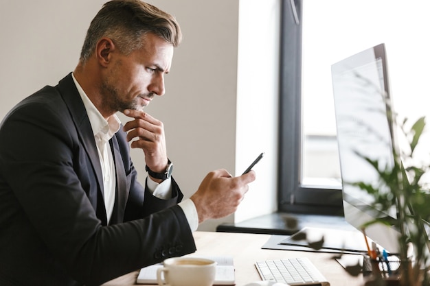 Portrait of unshaved businessman 30s wearing suit using mobile phone while working on computer in office