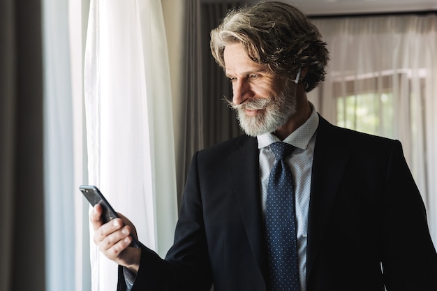 Portrait of unshaven adult businessman wearing black suit using smartphone and earbuds while standing near window in hotel apartment