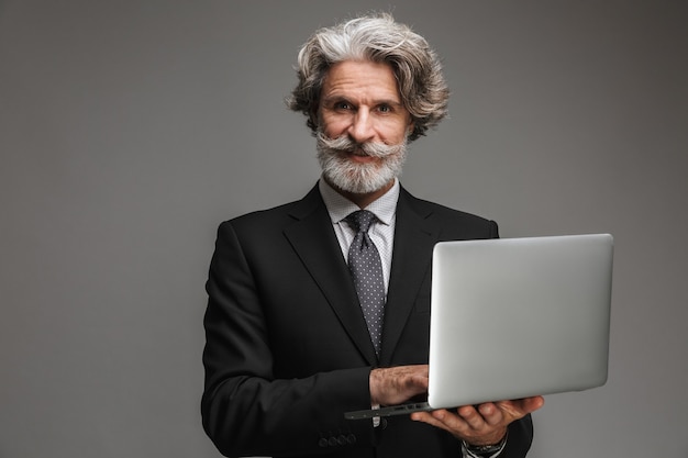 Portrait of unshaven adult businessman wearing formal black suit holding silver laptop isolated over gray wall