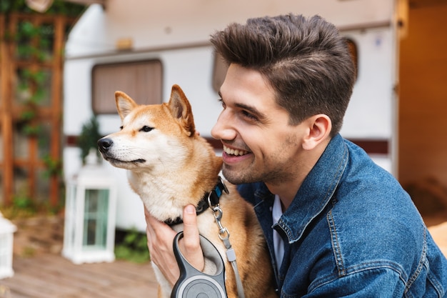 Portrait of unshaven smiling man wearing denim clothes hugging red dog near house on wheels outdoors