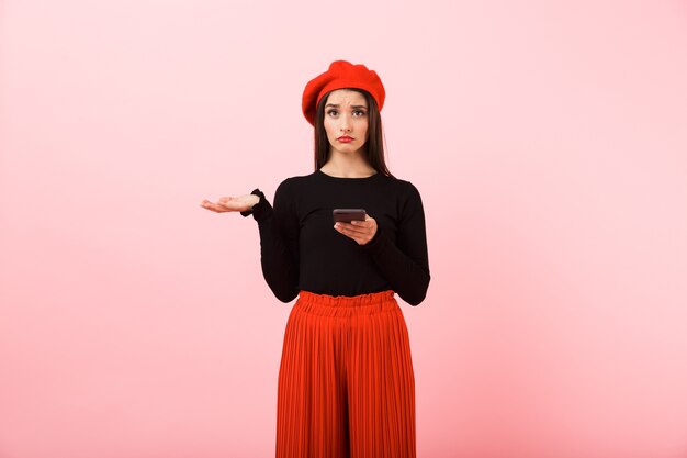 Portrait of an upset beautiful young woman wearing red beret standing isolated over pink background, holding mobile phone