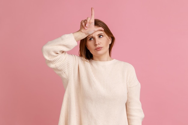 Portrait of upset blond woman standing with hand on forehead showing loser gesture unemployed or fired from job wearing white sweater Indoor studio shot isolated on pink background