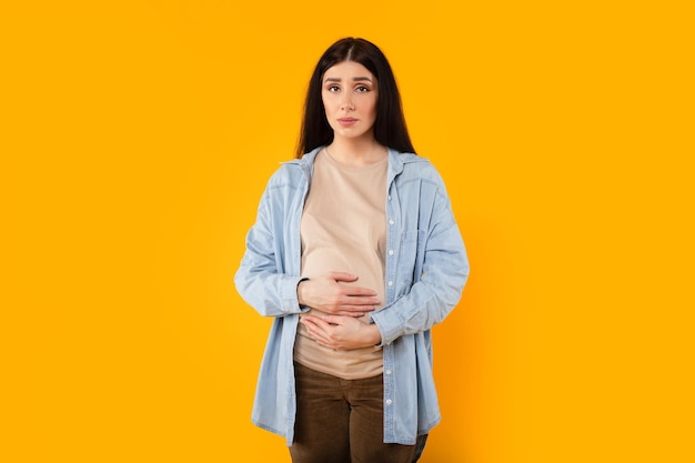 Portrait of upset caucasian pregnant woman touching belly and looking at camera standing over yellow