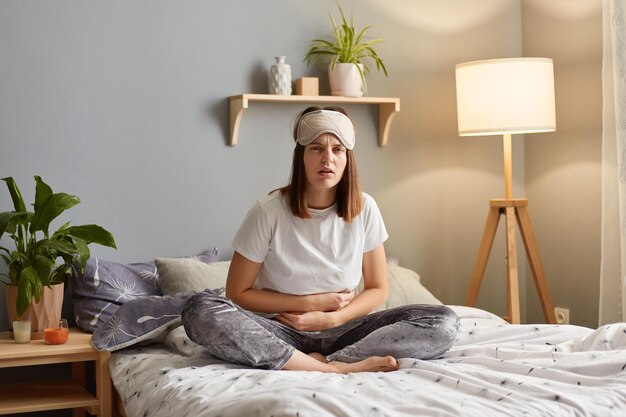 Portrait of upset Caucasian woman suffering from strong abdominal pain touching her tummy sitting on bed feeling acute stomachache has chronic disease spasm or hernia