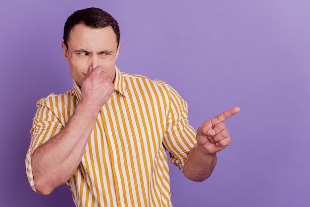 Portrait of upset disgusted guy direct forefinger empty space cover nose on purple background