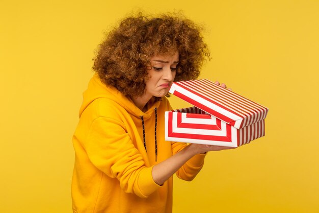 Portrait of upset displeased curlyhaired woman in hoodie looking into gift box opening present and peeking inside with dissatisfied expression indoor studio shot isolated on yellow background