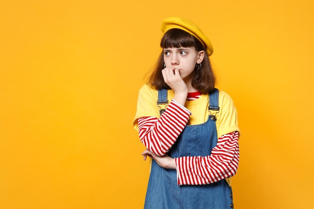 Ritratto di ragazza adolescente sconvolta in berretto francese, prendisole in denim che rosicchia le unghie guardando da parte isolato su sfondo giallo muro in studio. persone sincere emozioni concetto di stile di vita. mock up spazio di copia.