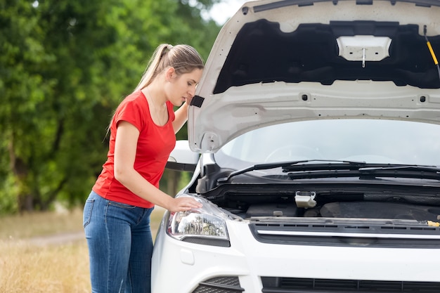 過熱した車のボンネットの下を見ている動揺した女性の肖像画