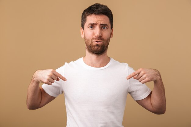 Portrait of an upset young man casually dressed standing isolated over beige, pointing fingers at his blank t-shirt