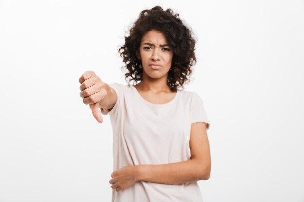 Portrait of upset young woman in basic t-shirt  and showing thumb down, isolated over white wall