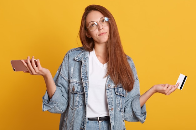 Portrait of upset young woman wears denim jacket and glasses, spreads hands, holds credit bank card and mobile phone, isolated on yellow wall. People lifestyle and online shopping concept.