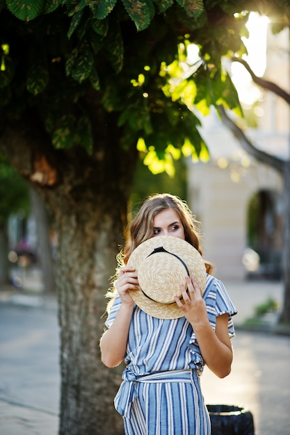 Portrait of a very attractive young woman in striped overall posing with her hat on a pavement in a town