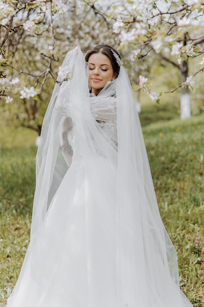 portrait of a very beautiful bride with long curly hair and wearing a white dress and long veil in a blooming spring garden Naarechena holds her veil with her hands on a green background of grass