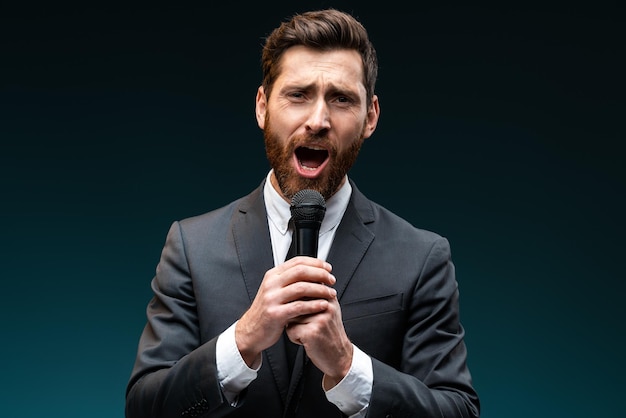 Portrait view positive bearded man in formal suit loudly singing song while holding microphone in hand. Man having fun resting in karaoke, singer performance