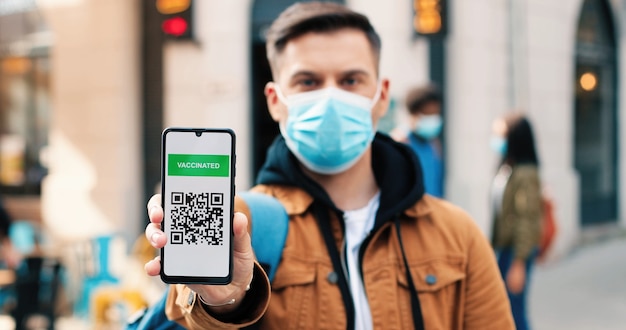 Portrait view of serious man wearing protective mask looking to camera and showing vaccine passport ...