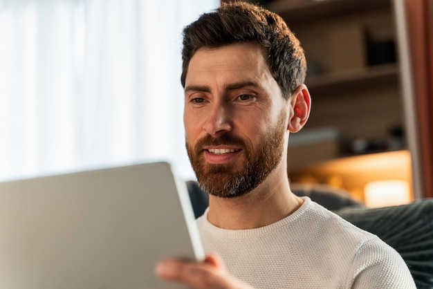 ベッドに座ってタブレット画面を見ている若い男の肖像画。彼はソーシャルネットワーキングで友達とチャットしながらタブレットを手に持っています